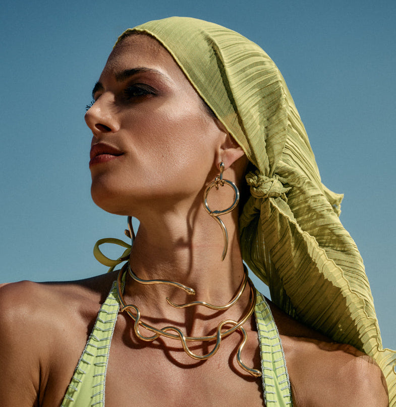 A woman outdoors wearing a bright yellow-green halter swimsuit with a matching pleated headscarf, layered Ellina's Treasures brand jewelry gold necklaces, and hoop earrings against a blue sky and rocky desert background.